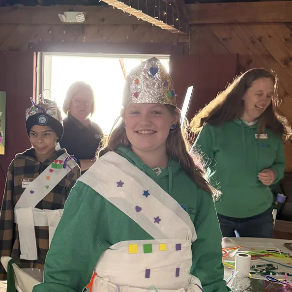 A camper wearing a foil crown and a paper costume she decorated herself, smiling in the craft lodge with DFC staff behind her