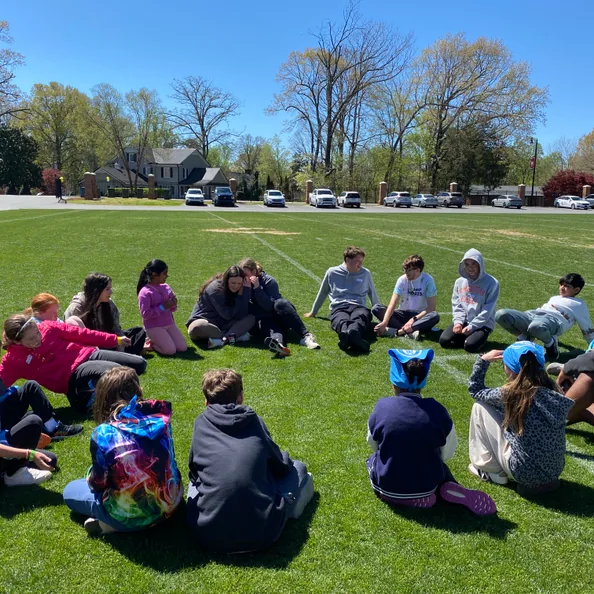 A wide circle of campers sitting cross-legged on the grass of a sports field on a clear spring day