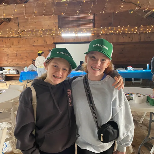 Two campers in matching green Dexcom caps standing arm in arm in the lodge, both grinning at the camera