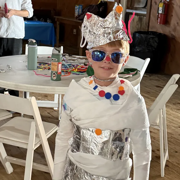 A younger camper in sunglasses and a costume of foil and tape, standing by the craft tables under the lodge string lights