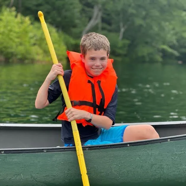 A camper in an orange life jacket paddling a canoe across the lake