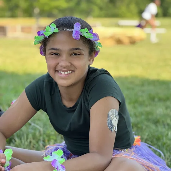 A Camp Morris camper in a flower crown sitting on the grass at luau night, a continuous glucose monitor on her arm