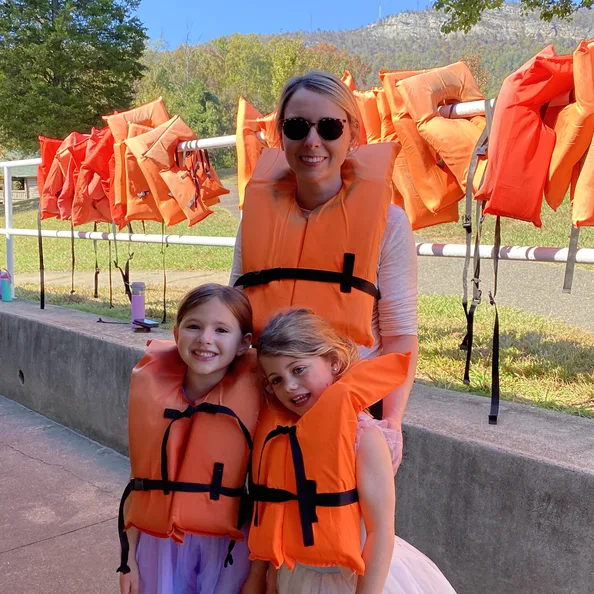 An adult and two children in orange life jackets at the lakefront, with rows of jackets hanging on the rack behind them