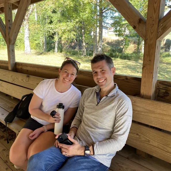 Two parents sitting together on a bench in the open-sided pavilion, sunlit woods behind them