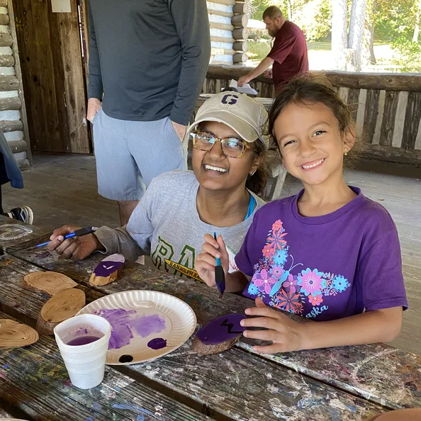 Two girls painting rocks at a picnic table in the craft cabin, a plate of purple paint between them