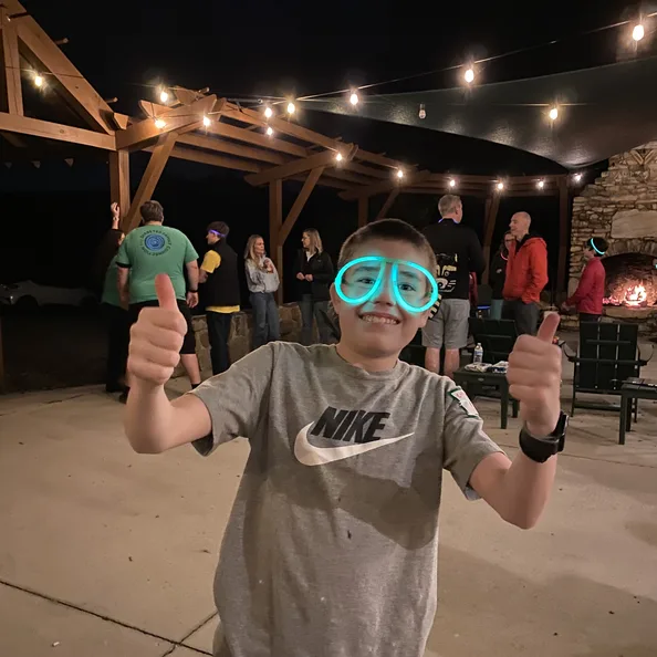 A boy giving two thumbs up at the evening gathering under the string lights, glow goggles pushed up on his forehead