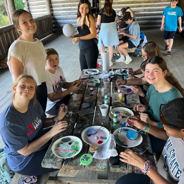 Teens painting rocks around a picnic table in the open-sided craft cabin, several of them looking up at the camera