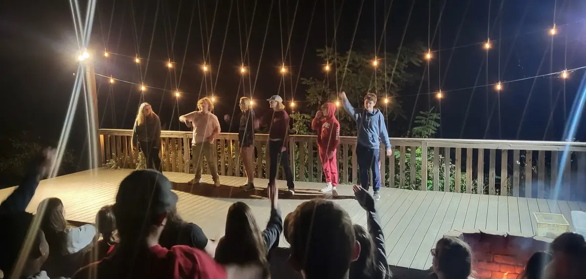 Teens performing on a deck strung with lights in the evening while the group watches