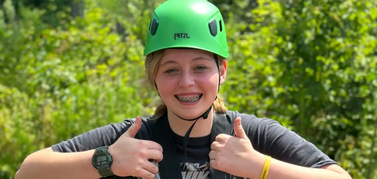 A teen in a green climbing helmet and harness giving two thumbs up before a climb