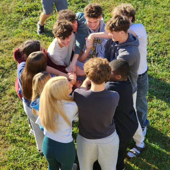 A dozen teens standing in a huddle on the lawn with their hands stacked in the middle, seen from above