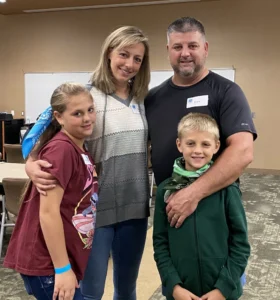 Two caregivers and their two children standing together at a Camp to Community event, the younger child wearing a green DFC bandana