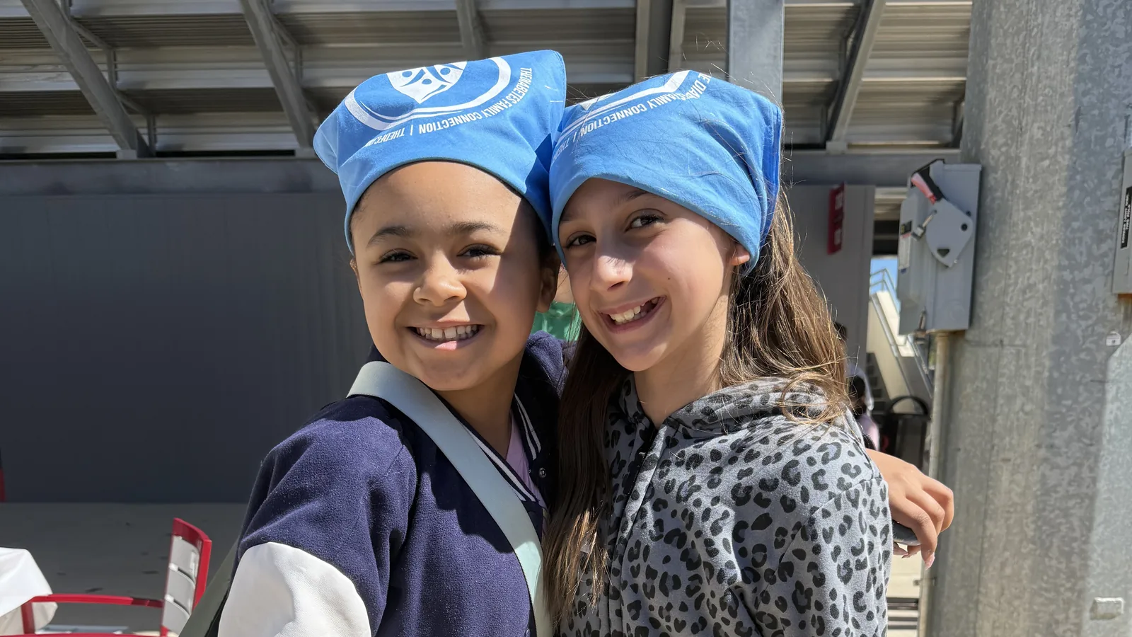 Two young campers in DFC bandanas stand arm in arm, smiling, at a Camp to Community day camp