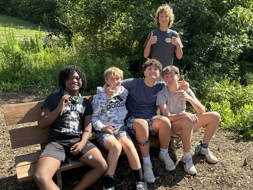 Five campers sitting together on a bench outdoors, smiling and giving thumbs up
