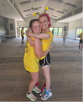 Iz and Aarlayn, counselors in yellow shirts and bee antennae, hugging in the dining hall