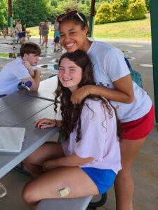 Kennedy hugging a fellow camper at a picnic table under the pavilion, a glucose monitor on the leg of the camper in front