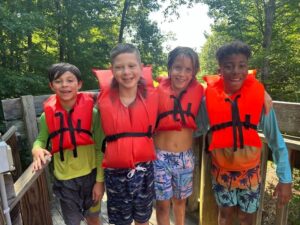 Four Camp Morris campers in orange life jackets standing arm in arm on a wooden bridge