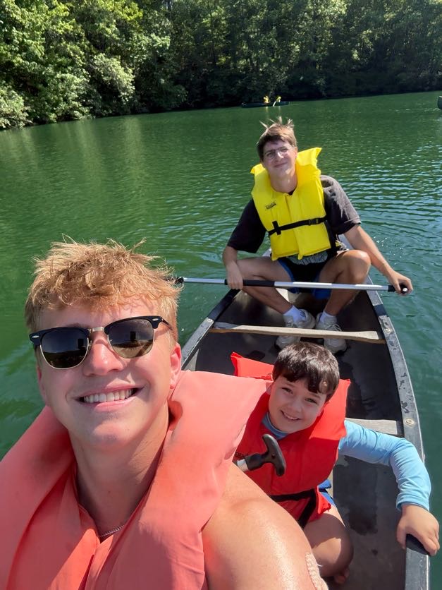 A counselor in sunglasses taking a selfie in a canoe with two campers in life jackets on the lake
