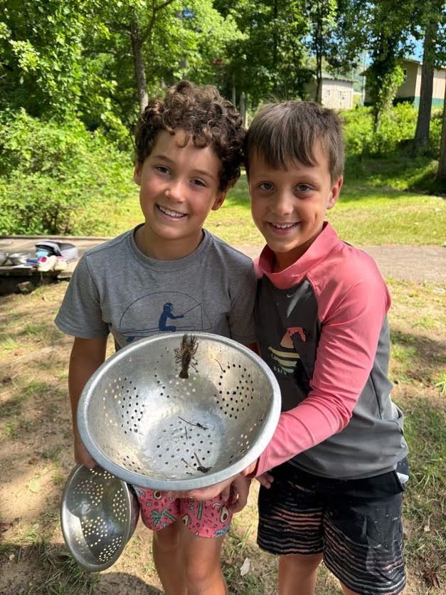 Two campers grinning as they hold up a colander with a crayfish they caught