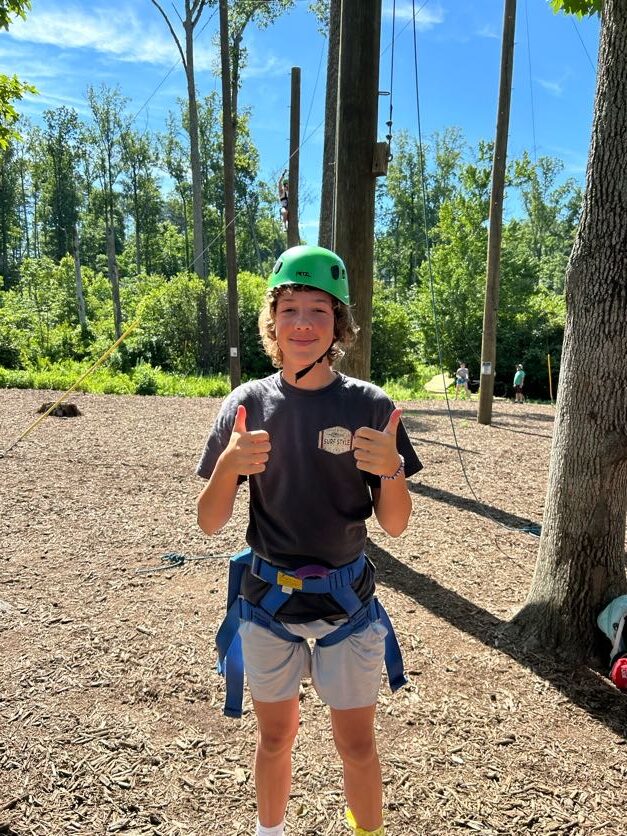 A camper in a green helmet and climbing harness giving two thumbs up on the ropes course
