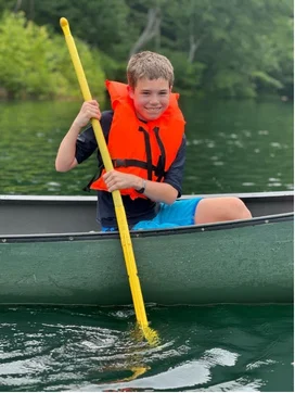 A smiling boy in an orange life jacket paddling a green canoe on the lake at the Family Retreat